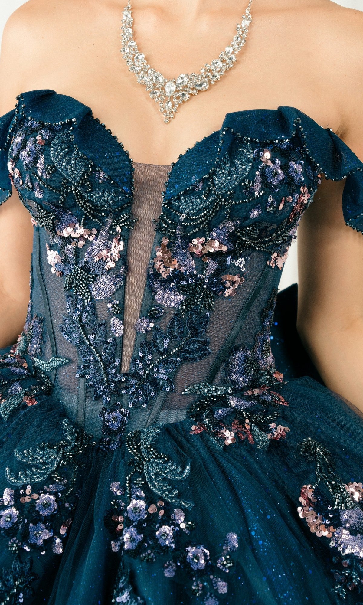 Close-up of a person wearing the Quinceanera Dress 8440JQ by Cinderella Couture—a dark blue sequined off-the-shoulder gown with floral beading, paired with a silver necklace.